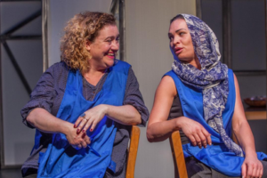 Photo of two women, both wearing blue aprons, sitting down facing each other