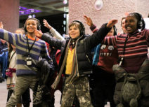 Photo of a group of children wearing headphones and standing with their arms outstretched, smiling