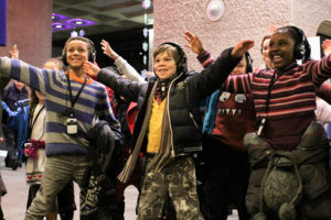 Photo of a group of children wearing headphones and standing with their arms outstretched, smiling
