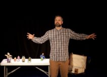 Man in checked shirt standing with his arms outstretched in front of a trestle table with small figurines on it