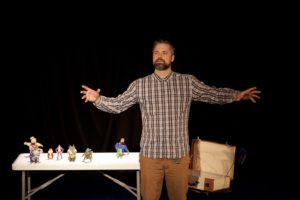 Man in checked shirt standing with his arms outstretched in front of a trestle table with small figurines on it