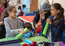 A family are gathered around a table doing art activities together. One girl is holding a black, cardboard tube up to her eye and looking through it.