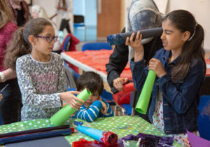 A family are gathered around a table doing art activities together. One girl is holding a black, cardboard tube up to her eye and looking through it.