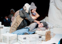 Production image for 'Where's My Igloo Gone?'. A woman wearing a grey hat and jumper using a blue coat as a blanket is leaning on white coloured wooden crates. Another woman wearing a grey beanie hat and beige and black coat and is leaning over her as though trying to wake her up.