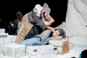 Production image for 'Where's My Igloo Gone?'. A woman wearing a grey hat and jumper using a blue coat as a blanket is leaning on white coloured wooden crates. Another woman wearing a grey beanie hat and beige and black coat and is leaning over her as though trying to wake her up.
