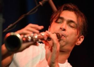 A close-up photo of a man with dark hair and a white t-shirt playing a clarinet.