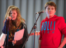 Photo of a teenage girl and boy. The girl is stood on the left and is wearing a navy jumper, she has long blonde hair. The boy is wearing a red 'Hollister' jumper and has short brown hair. Both children are standing behind microphones.