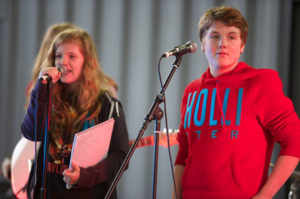 Photo of a teenage girl and boy. The girl is stood on the left and is wearing a navy jumper, she has long blonde hair. The boy is wearing a red 'Hollister' jumper and has short brown hair. Both children are standing behind microphones.