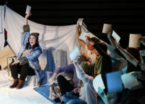 Production photo of Where's My Igloo Gone? An actor in a blue coat is holding up a piece of paper, the audience is sat to her right, looking at her and also holding up pieces of paper.