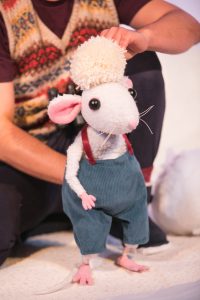Production image from Snow Mouse. A white toy mouse with pink hands, ears and feet is wearing blue dungarees. A performer is operating the mouse like a puppet and is holding a cream pompom on his head.