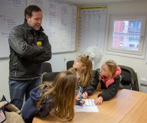 A man is standing in an office space in front of two whiteboards, three girls are sat looking up at him. They all have felt tip pens in their hands and their drawing is on a table in front of them.