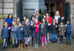 Photo of a group of approximately 12 children and 4 adults stood in front of Z-arts. They are all wearing coats and waving at the camera.