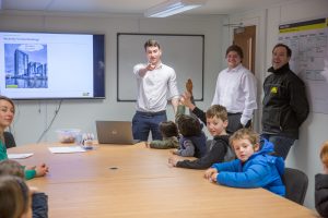 Photo of a man giving a powerpoint presentation to children sat around a boardroom style table.