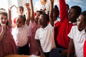 Picture of school children. The girls in the photo are wearing red checked summer dresses and the boys are wearing white polo shirts or red jumpers. Most of the children have a hand raised in the air as if wanting to ask or answer a question.