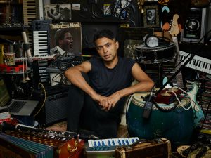 A man is sat on the floor surrounded by different musical instruments including drums, keyboards, accordians, guitars and musical speakers, headphones and amps.
