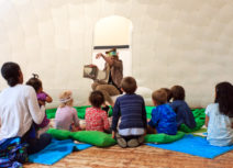 A woman dressed in animal print is sitting in front of a group of children reading a story to them. The children are sat on green cushions and blue mats.