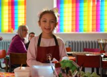Picture of child smiling directly at the camera, she is wearing a pink jumper and a red apron. In the foreground are paint brushes, egg boxes and other arts and crafts supplies. In the background are rainbow coloured window blinds.