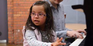 A girl approximately 6 years old is sitting at a piano. She is wearing a grey polkadot jumper and a light pink stripey gilet. Next to her is an adult female wearing a black and white stripey shirt