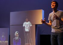 Production image from Friends For All. A man is standing in front of projected images of children who are wearing school uniform, one child is stood on a table wearing a white t-shirt that says 'fight the power' on it),