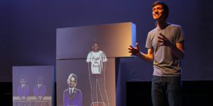 Production image from Friends For All. A man is standing in front of projected images of children who are wearing school uniform, one child is stood on a table wearing a white t-shirt that says 'fight the power' on it),