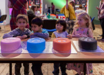 Four children stood behind a table with four cakes, one pink, one blue, one orange and one black.