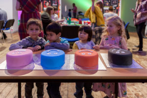 Four children stood behind a table with four cakes, one pink, one blue, one orange and one black.