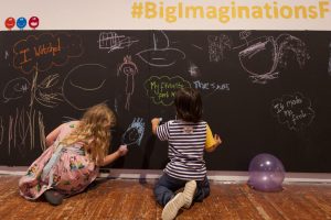 Picture of two children drawing on a chalk-board painted wall, one is wearing a pink, floral patterned dress and one is wearing a white and blue striped t-shirt with jeans.