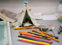 Picture of a small teepee tent made of push pins, beige fabric and pencils. The toy tent is behind a pile of coloured pencil crayons.