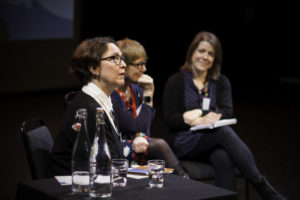 Photo of three women seated. They are all wearing dark colours. In the foreground are two bottles of water and two glasses.