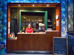 A staff member wearing a red Z-arts polo shirt stands behind the wooden counter at Z-cafe, In the background is a coffee machine, jars with different types of teas, wine and pint glasses, a selection of fruit and various other food items.