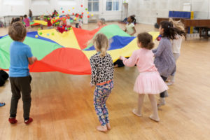 Lead image for Visiting Z-arts page. Picture of a group of 4 - 6 year olds standing in a circle holding a multi-coloured parachute. There are small multi-coloured balls bouncing on the parachute and the children are stood on a wooden, laminate floor.