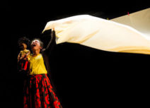 Woman wearing a yellow top and black and red patterned shirt is waving a white sheet.