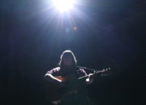 Photo of a woman holding a guitar. She is standing under a spotlight and silhouetted by the light.