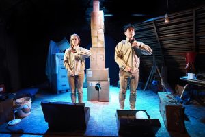 One Small Step production image. A man and a woman dressed in beige are facing forward, zipping up their jackets in unison. In the background is a tower made of cardboard boxes.