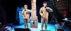 One Small Step production image. A man and a woman dressed in beige are facing forward, zipping up their jackets in unison. In the background is a tower made of cardboard boxes.