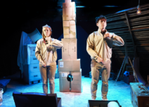 One Small Step production image. A man and a woman dressed in beige are facing forward, zipping up their jackets in unison. In the background is a tower made of cardboard boxes.