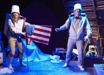 Production shot - One Small Step. A man and a woman dressed in beige are wearing buckets on their heads. The woman is holding an american flag and they are both wearing cardboard boxes as though they are rucksacks