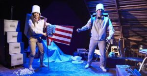 Production shot - One Small Step. A man and a woman dressed in beige are wearing buckets on their heads. The woman is holding an american flag and they are both wearing cardboard boxes as though they are rucksacks