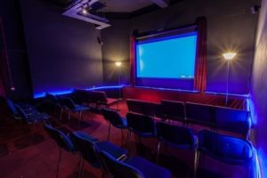 Photo of Z-arts cinema room. A blue screen is at the front of the room with red curtains drawn at either side, there is a lamp to either side of the screen. The room has a red carpet and the are three rows of seats with an aisle down the middle.