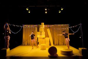 Production image from Tidy Up, three dancers wearing black tshirts and bronze shorts stand on a set with yellow curtains, fairy lights and a tower of large yellow sponge shapes.