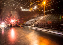 Photo of the interior of Z-arts theatre space. The stage is flat with a black, laminate floor. There is a raked seating bank of 250 black theatre seats with an aisle of steps down the middle.