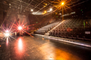 Photo of the interior of Z-arts theatre space. The stage is flat with a black, laminate floor. There is a raked seating bank of 250 black theatre seats with an aisle of steps down the middle.
