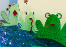 Picture of green paper frogs with red tongues and big eyes made by children and families as part of Family Art Stars. The frogs are arranged on a piece of shiny, sparkly, blue paper.