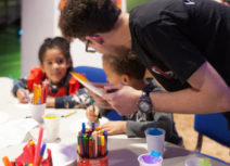 A man and two children doing art activities. The man is stood up and the two children are sitting down at a table. On the table are felt tip pens, pencil crayons, pieces of paper and plastic cups filled with coloured tissue paper.