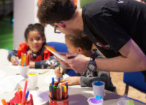 A man and two children doing art activities. The man is stood up and the two children are sitting down at a table. On the table are felt tip pens, pencil crayons, pieces of paper and plastic cups filled with coloured tissue paper.