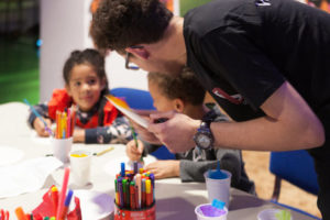 A man and two children doing art activities. The man is stood up and the two children are sitting down at a table. On the table are felt tip pens, pencil crayons, pieces of paper and plastic cups filled with coloured tissue paper.