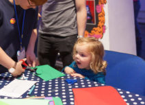 A young blonde girl sits at a table covered with a navy blue and white polkadot table-cloth. An adult woman standing next to her cutting a shape out of a piece of green paper.