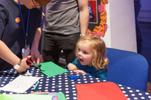 A young blonde girl sits at a table covered with a navy blue and white polkadot table-cloth. An adult woman standing next to her cutting a shape out of a piece of green paper.