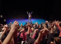 Picture of Comedy Club 4 Kids audience - a man stands on stage with his arms raised in front of an audience of children who all have their arms raised in the air.