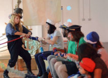 A woman wearing a top hat and black dungarees hands a cardboard anchor to a woman sitting down wearing a sailor hat. Sitting next to her are two men, two women and a child all wearing hats.